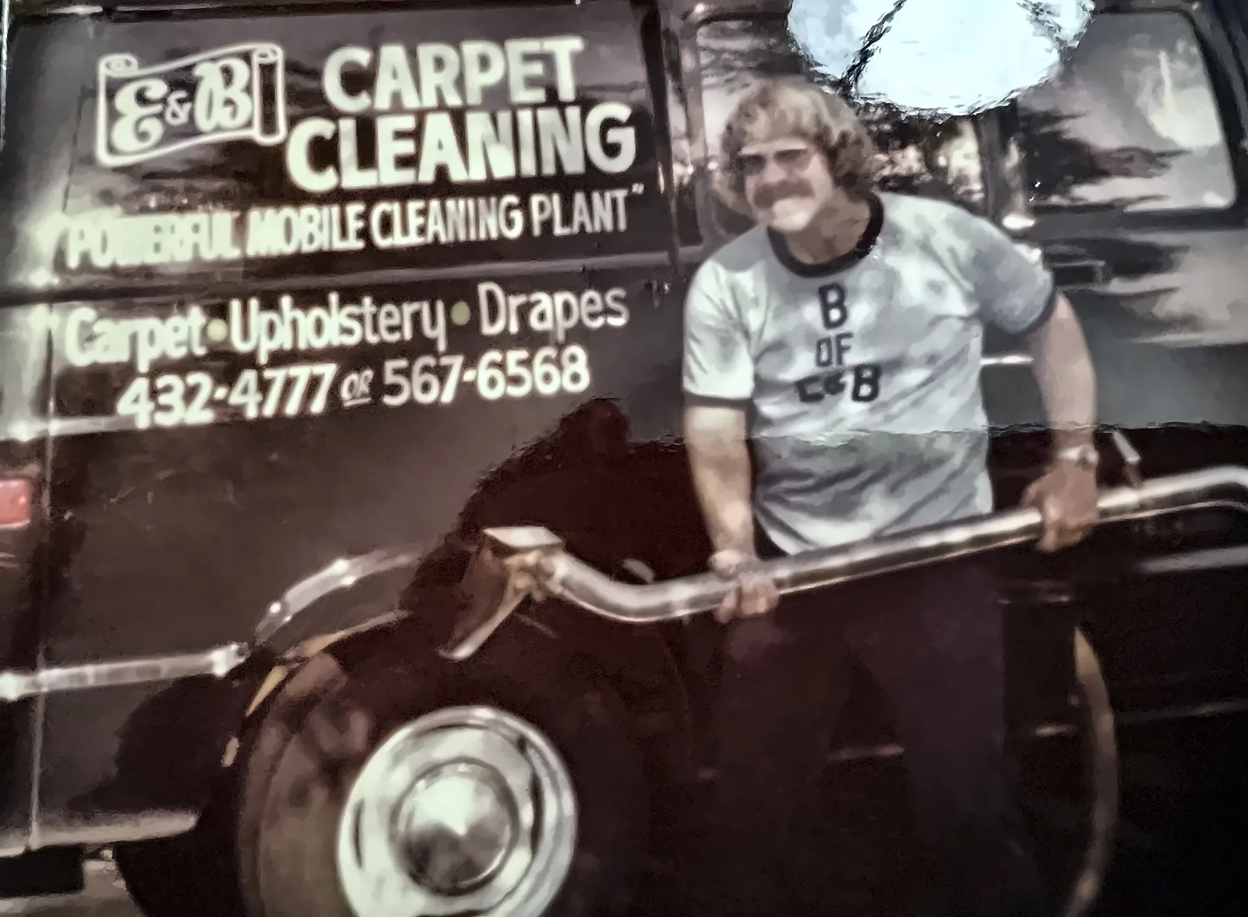 Brian, the B of E&B, in an E&B T-shirt holding a cleaning wand beside the black E&B Carpet Cleaning van, 1970s